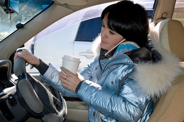 Giovane donna alla guida dell'auto, bere caffè e parlare al telefono
