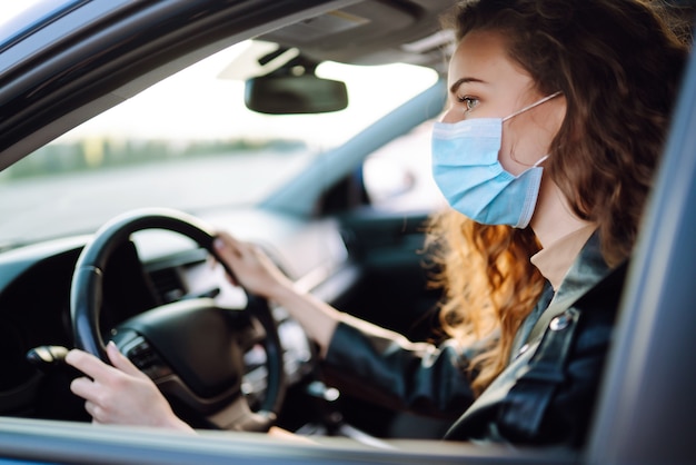 Giovane donna alla guida di un'auto che indossa una maschera medica durante un'epidemia. Isolamento del trasporto. Covid-2019.