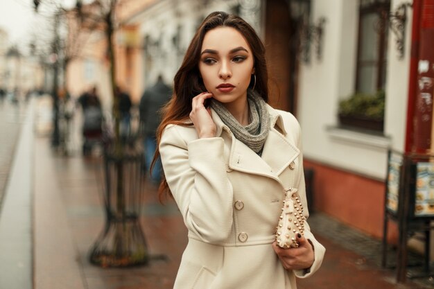 Giovane donna alla moda alla moda in un cappotto di autunno alla moda con una borsa alla moda che cammina all'aperto in città