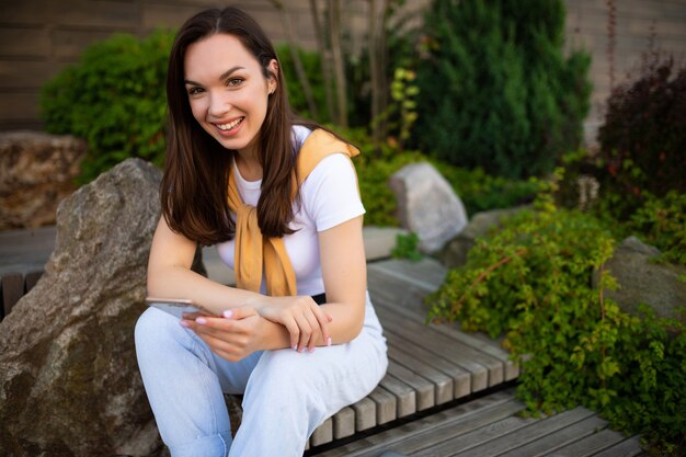 Giovane donna alla moda che gode del tempo libero che si siede nel parco verde di estate e che chiacchiera sul telefono.