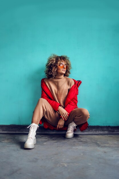 Giovane donna alla moda con i capelli ricci, in giacca rossa accovacciata davanti al muro e guardando lontano.