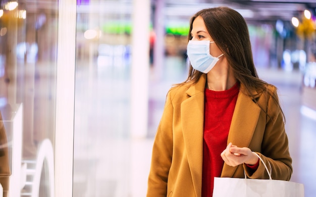 Giovane donna alla moda fiduciosa in una mascherina medica di sicurezza con una borsa della spesa sta camminando nel centro commerciale