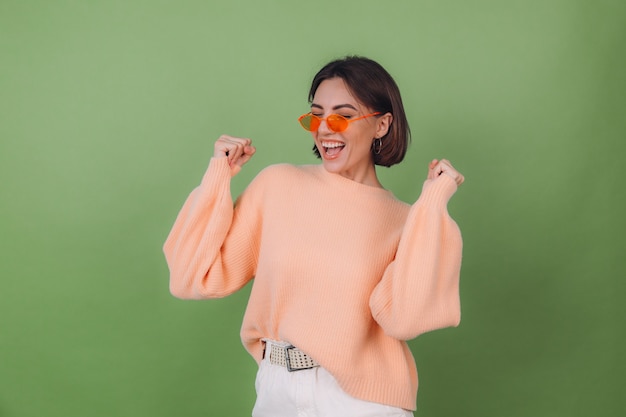 Giovane donna alla moda in maglione pesca casual e occhiali arancioni isolati su muro verde oliva positivo facendo gesto vincitore pugno di serraggio spazio copia