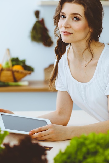 Giovane donna alla ricerca di una nuova ricetta per cucinare in cucina. La casalinga sta facendo acquisti online tramite tablet e carta di credito.