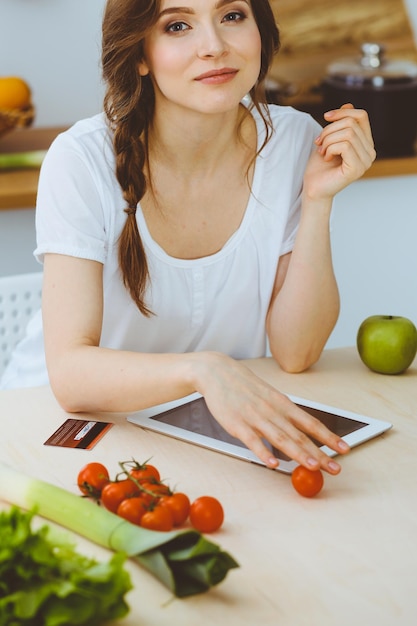 Giovane donna alla ricerca di una nuova ricetta per cucinare in cucina. La casalinga sta facendo acquisti online tramite tablet e carta di credito.