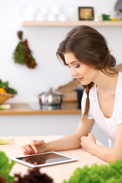 Giovane donna alla ricerca di una nuova ricetta per cucinare in cucina. La casalinga sta facendo acquisti online tramite tablet e carta di credito