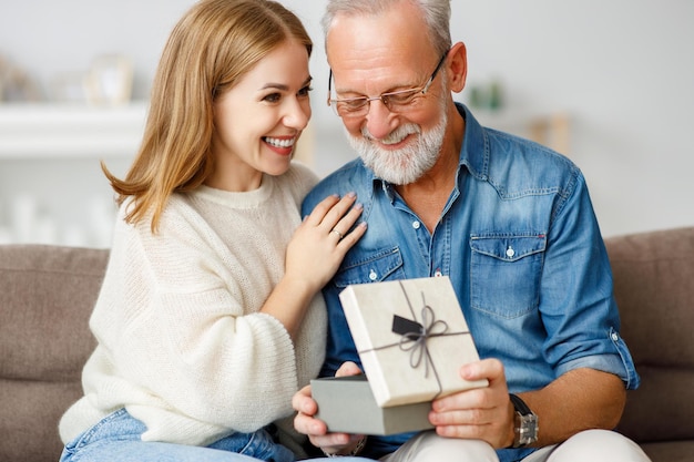 Giovane donna allegra che abbraccia l'uomo anziano mentre è seduto sul divano e apre la scatola del regalo durante la celebrazione delle vacanze a casa