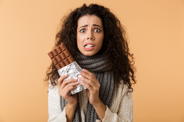 Giovane donna allegra che indossa un maglione e sciarpa che tiene grande barra di cioccolato isolata sopra la parete beige