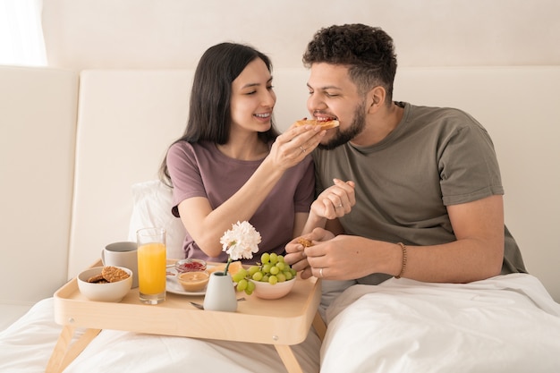 Giovane donna allegra che mette un panino con marmellata nella bocca del marito mentre entrambi sono seduti nel letto matrimoniale e si godono la colazione al mattino