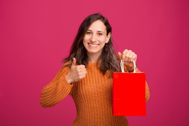 Giovane donna allegra che porta maglione giallo che tiene il sacchetto della spesa rosso e che mostra i pollici su