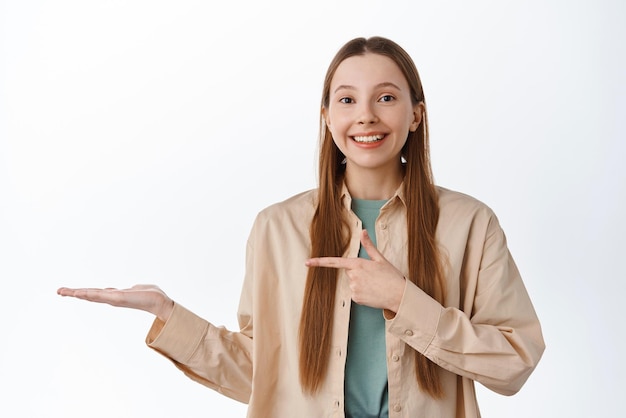Giovane donna allegra che punta il dito a mano aperta che mostra il prodotto dimostra l'elemento promozionale sul palmo contro il copyspace visualizza il tuo logo in piedi su sfondo bianco