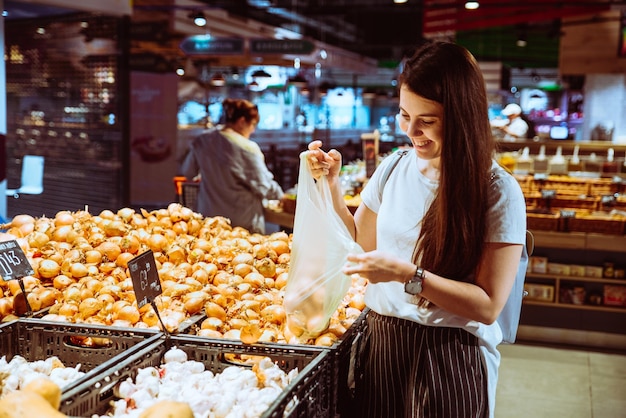 Giovane donna allegra che sceglie le cipolle nel concetto di acquisto di drogheria del negozio