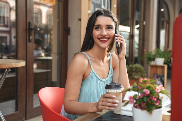 giovane donna allegra che si siede nella caffetteria all'aperto mentre parla dal telefono cellulare bere caffè