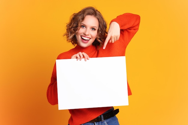 Giovane donna allegra che sorride per la macchina fotografica e che punta a banner vuoto su sfondo giallo