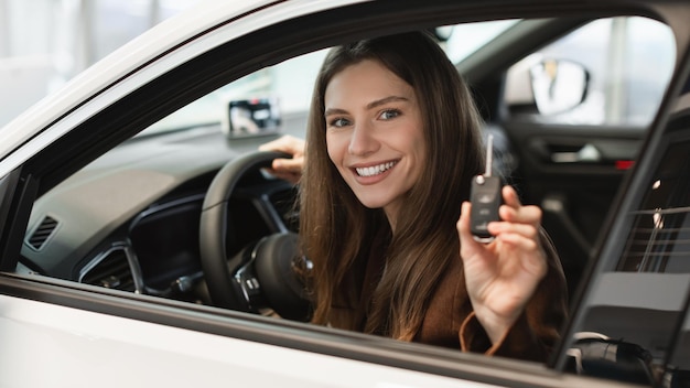 Giovane donna allegra che tiene la chiave dall'auto che acquista una nuova auto allo showroom seduto all'interno dell'automobile