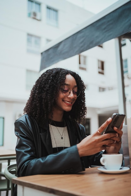 Giovane donna allegra che usa il suo smartphone mentre sta bevendo un caffè sulla terrazza di un caffè