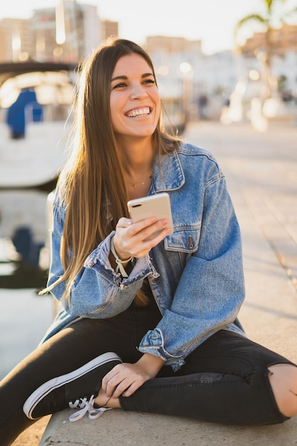 Giovane donna allegra che usa il telefono mentre è seduta al porto durante il tramonto