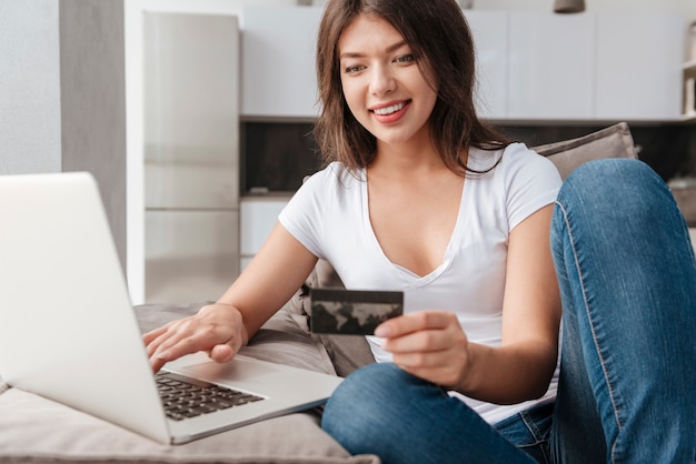 Giovane donna allegra che utilizza laptop e carta di credito sul divano di casa