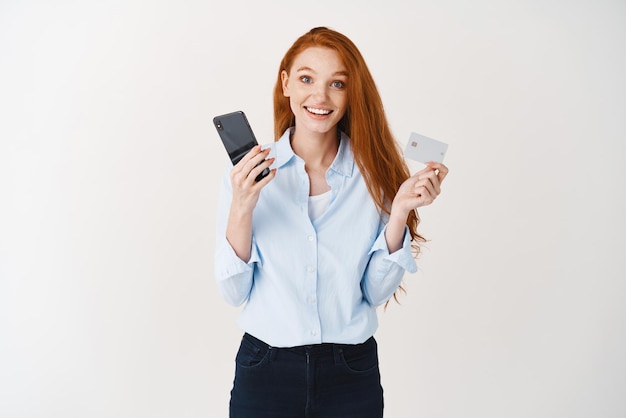 Giovane donna allegra con i capelli rossi che compera online che tiene la carta di credito di plastica con il telefono cellulare e che sorride felice alla priorità bassa bianca della macchina fotografica