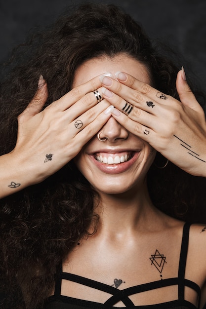 giovane donna allegra con un tatuaggio che sorride e si copre gli occhi isolati su un muro nero