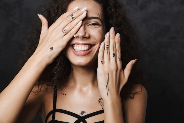 giovane donna allegra con un tatuaggio che sorride e si copre il viso isolato su un muro nero