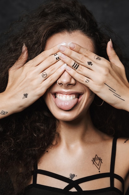 giovane donna allegra con un tatuaggio che sporge la lingua e si copre gli occhi isolati su un muro nero