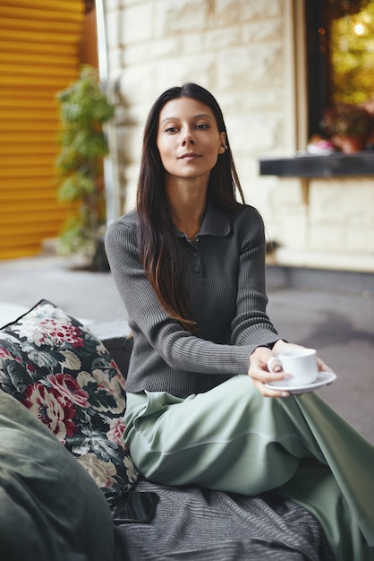 Giovane donna allegra con una tazza di caffè che si siede al caffè che sorride all'aperto