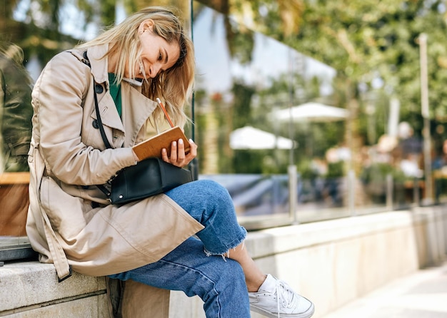 Giovane donna allegra e candida che sorride ampiamente mentre parla con un amico sullo smartphone seduto all'aperto in città in una giornata di sole Bella donna che parla sul cellulare scrivendo alcuni avvisi sul taccuino