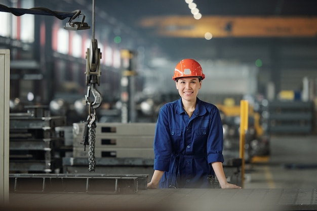 Giovane donna allegra in abbigliamento da lavoro e casco protettivo che ti guarda mentre lavora in un moderno impianto industriale