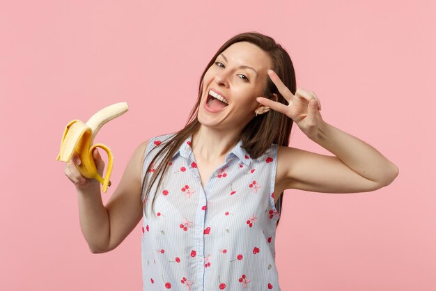 Giovane donna allegra in abiti estivi che mostrano il segno di vittoria che tiene frutta fresca di banana matura isolata su sfondo rosa pastello. Persone stile di vita vivido, relax concetto di vacanza. Mock up spazio di copia.