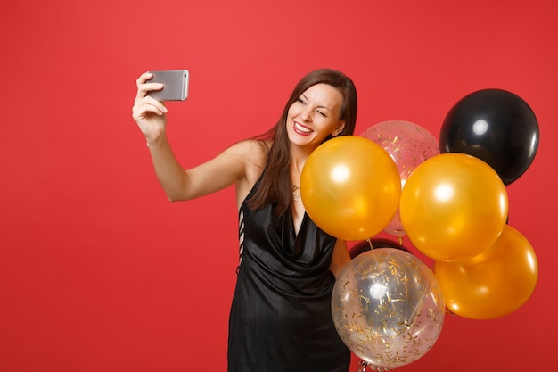 Giovane donna allegra in abito nero che celebra tenendo gli aerostati di aria lampeggiante facendo prendendo selfie girato sul telefono cellulare isolato su sfondo rosso. Felice Anno Nuovo compleanno mockup festa concetto.