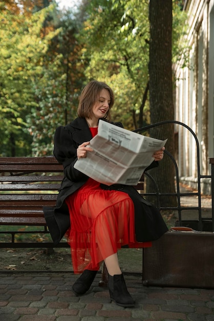 Giovane donna allegra in abito rosso e cappotto nero si siede sul banco leggendo il giornale e ridendo Pretty woman