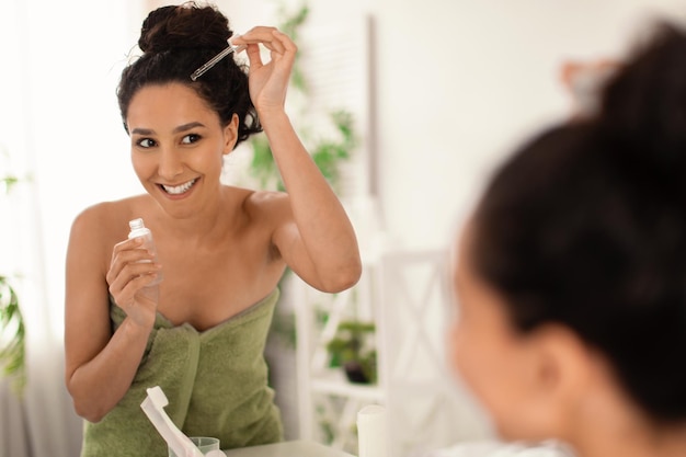 Giovane donna allegra in asciugamano che applica siero o olio naturale sui suoi capelli vicino allo specchio a casa copia spazio