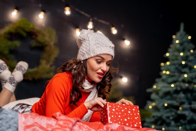 Giovane donna allegra in cappello bianco che esamina il presente prima di aprirlo Celebrazione del nuovo anno