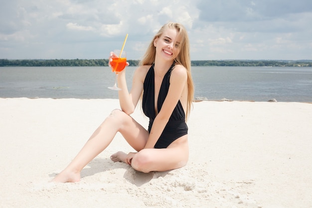 Giovane donna allegra in costume da bagno, brindando con un cocktail, bevendo bevande fresche sulla spiaggia, via mare. Concetto di vacanza.