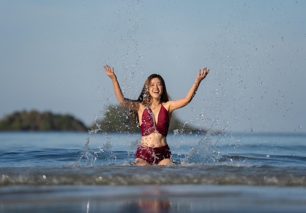 Giovane donna allegra in costume da bagno che gioca a spruzzi d'acqua sulla spiaggia del mare all'isola di Koh Chang Thailandia