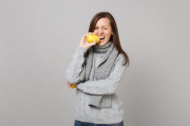 Giovane donna allegra in maglione grigio, sciarpa lampeggiante, limone mordace isolato su sfondo grigio in studio. Stile di vita sano, persone sincere emozioni, concetto di stagione fredda. Mock up copia spazio.