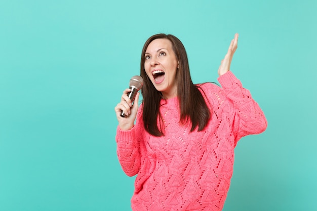 Giovane donna allegra in maglione rosa lavorato a maglia che balla, allarga le mani, tiene cantare una canzone nel microfono isolato su sfondo blu della parete, ritratto in studio. Concetto di stile di vita della gente. Mock up copia spazio.