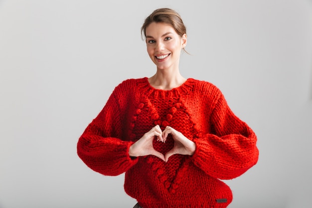 giovane donna allegra in maglione rosso che fa il gesto del cuore con le dita isolate su sfondo bianco