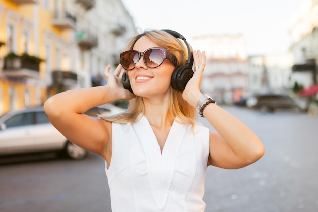 Giovane donna allegra in occhiali da sole che ascolta musica con le cuffie in città