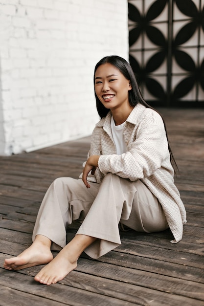 Giovane donna allegra in pantaloni beige e giacca a quadri oversize ride e si siede sul pavimento di legno su sfondo bianco muro di mattoni