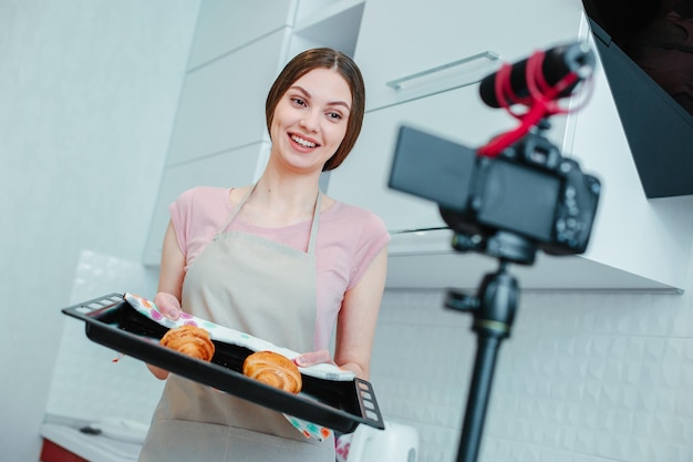 Giovane donna allegra in piedi in cucina davanti a una telecamera e sorridente mentre si tiene una teglia con pasticceria