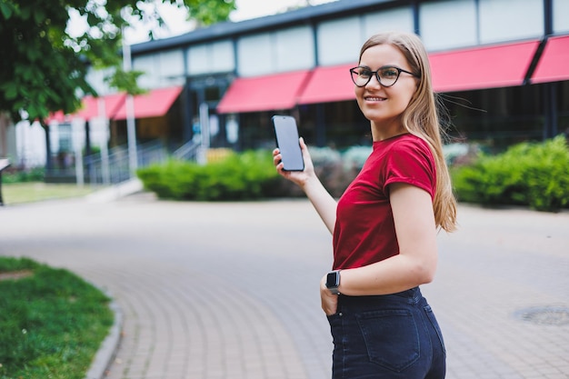 Giovane donna allegra in pullover grigio e blue jeans e occhiali in piedi sul marciapiede che trasporta borse della spesa e messaggi sullo smartphone a New York