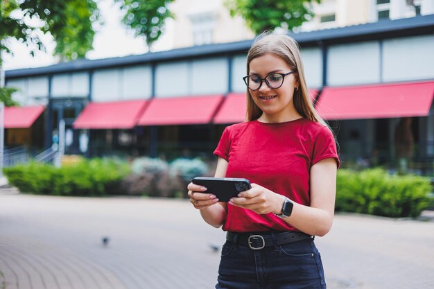 Giovane donna allegra in pullover grigio e blue jeans e occhiali in piedi sul marciapiede che trasporta borse della spesa e messaggi sullo smartphone a New York