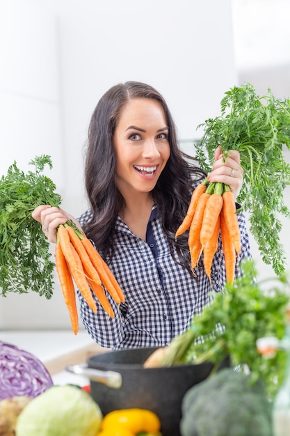 Giovane donna allegra nella sua cucina che tiene la carota fresca in entrambe le mani - concetto di verdura e di brughiera di dieta.