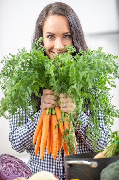 Giovane donna allegra nella sua cucina che tiene la carota fresca in entrambe le mani - concetto di verdura e di brughiera di dieta.