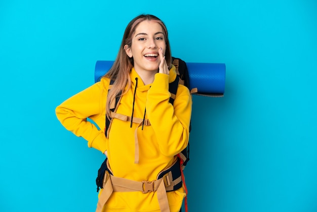 Giovane donna alpinista con un grande zaino isolato su blu che grida con la bocca spalancata