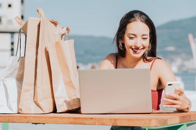 Giovane donna araba accanto ad alcune borse della spesa che fanno shopping online sul laptop, giornata dello shopping, stile moderno, sorridente durante una giornata di sole, shopping online