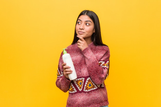 Giovane donna araba che tiene una bottiglia crema che guarda lateralmente con espressione dubbiosa e scettica.