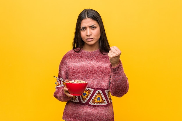 Giovane donna araba che tiene una ciotola di cereali che mostra il pugno alla macchina fotografica, espressione facciale aggressiva.